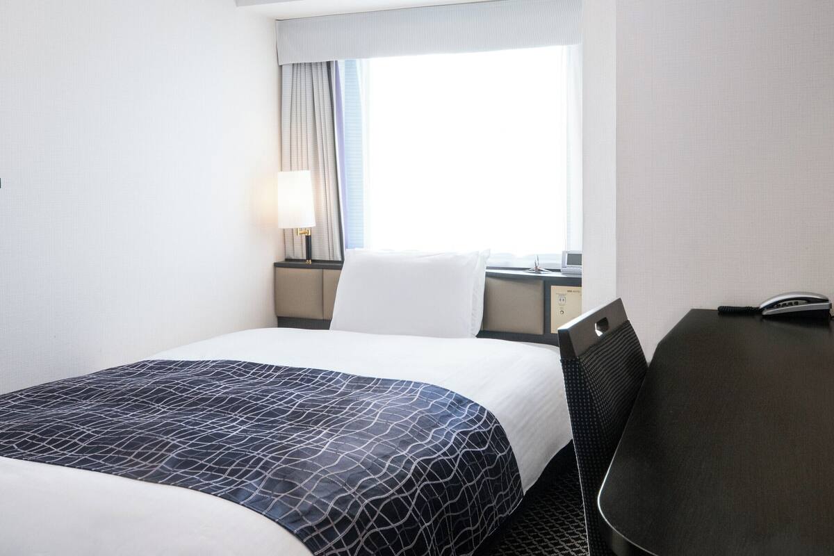 Een hotelkamer met een bed, een bureau met telefoon, een lamp en een raam met gordijnen.