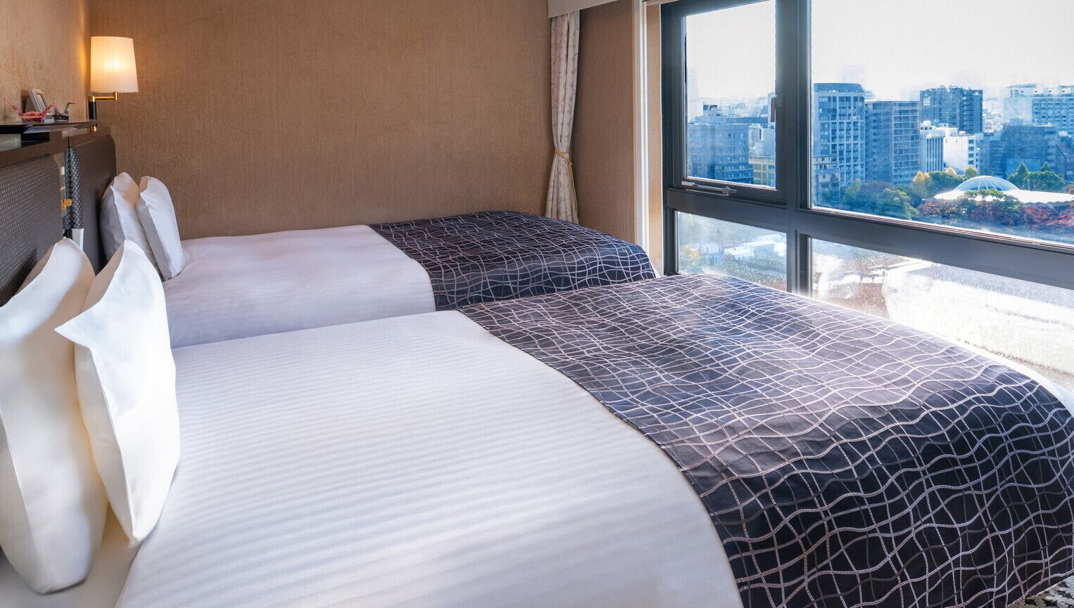 Une chambre d’hôtel avec deux lits, une vue sur la ville, une lampe et une fenêtre avec des rideaux.