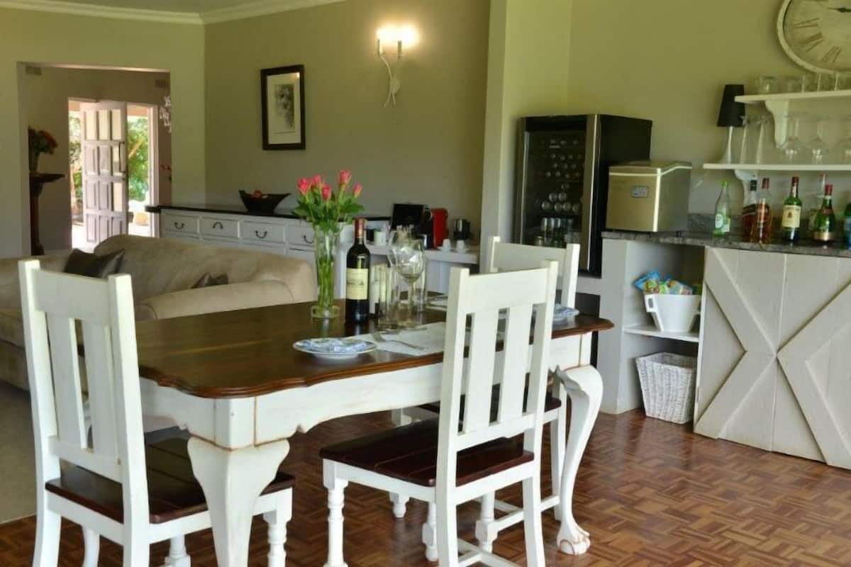 A dining area with a wooden table and white chairs, a kitchen with a refrigerator and shelves, and a door leading outside.
