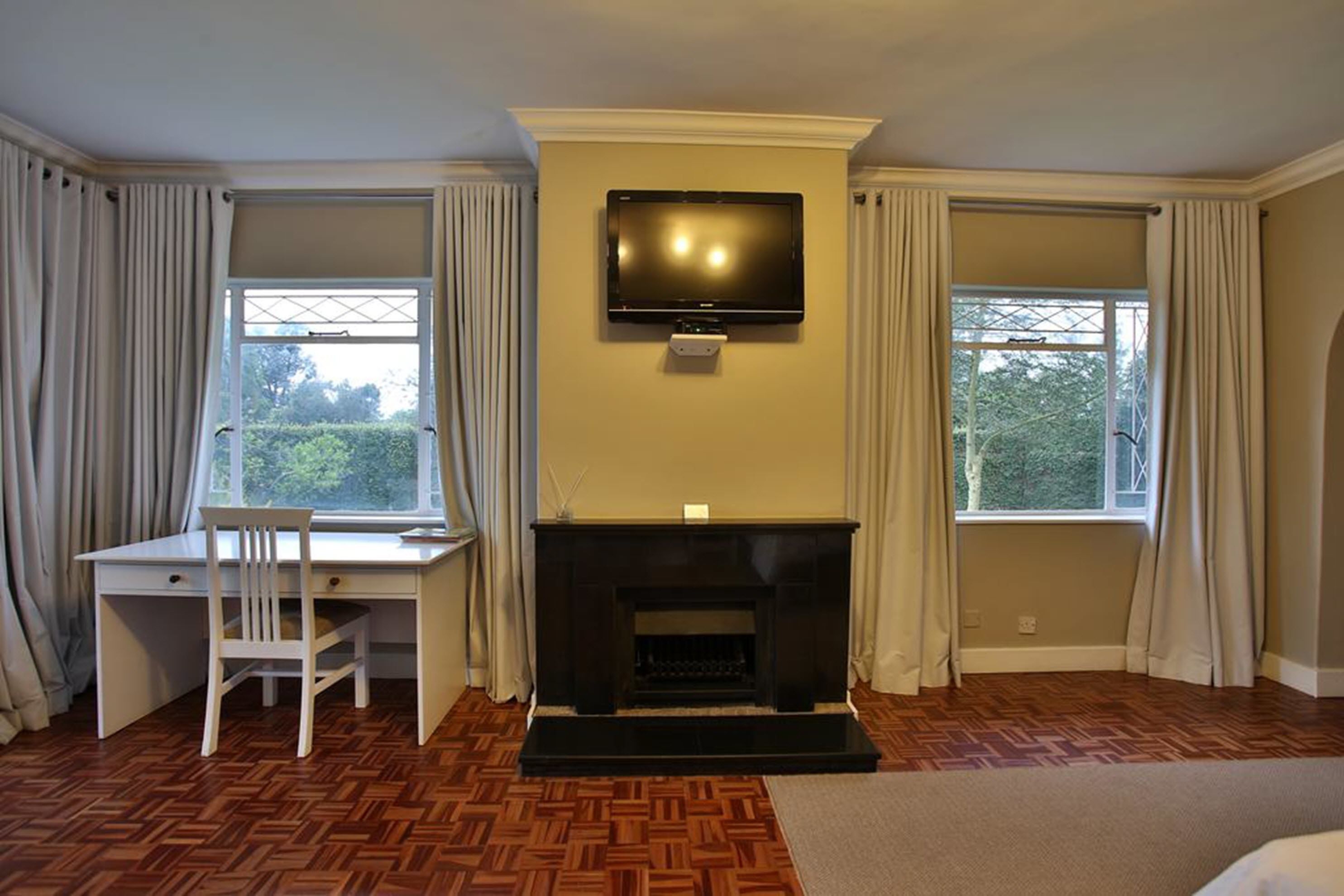 A room with a fireplace, a television mounted above it, a desk with a chair, and two windows with curtains.