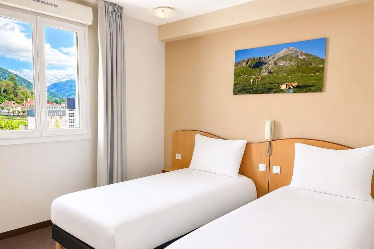 A hotel room with two beds, a window with a view of mountains and buildings, a painting of a mountain landscape, and a telephone on the wall.