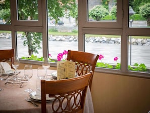 A dining table set with wine glasses and a menu, a wooden chair, and a view of a river and greenery through large windows.