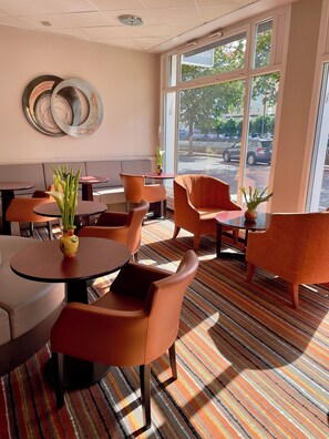 A modern indoor seating area with round tables and orange chairs, large windows with a view of the street, and decorative wall art. - Hotel Saint Georges Lourdes Pyrenees (Lourdes)