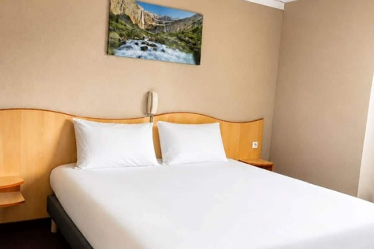 A hotel room with a bed, wooden headboard, white bedding, a nightstand, a telephone, and a painting of a mountain landscape.