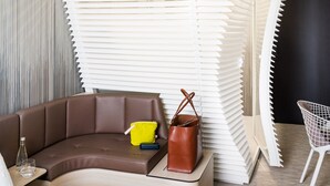 Una habitación de hotel moderna con un sofá marrón curvo, una mesita con un vaso, un bolso amarillo y un bolso de cuero marrón.