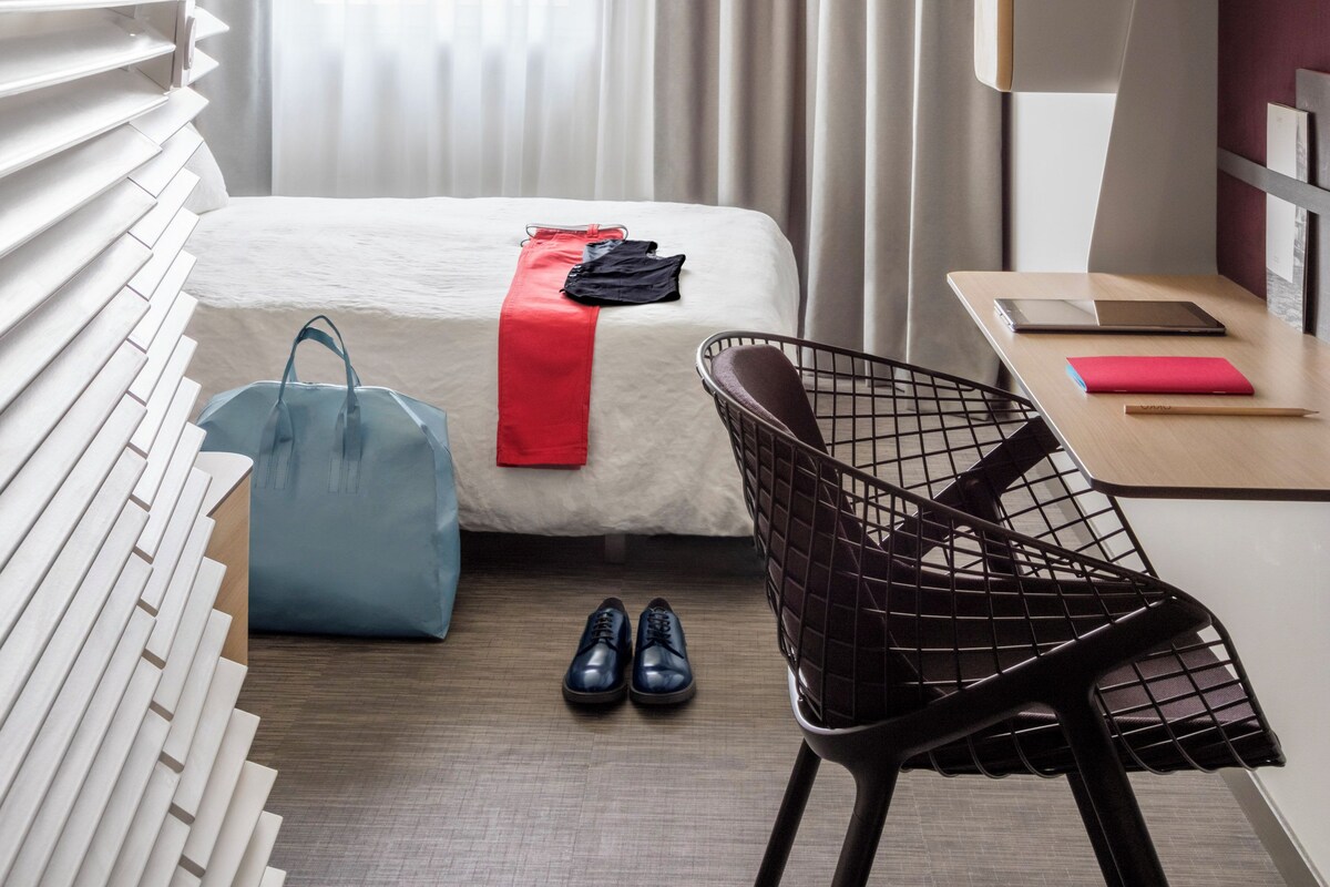 Une chambre d’hôtel moderne équipée d’un lit, d’un bureau, d’une chaise, d’une valise et d’une paire de chaussures.