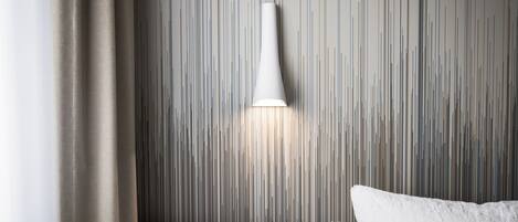 Una habitación de hotel moderna con pared texturizada, una lámpara blanca y una cama con ropa de cama blanca.
