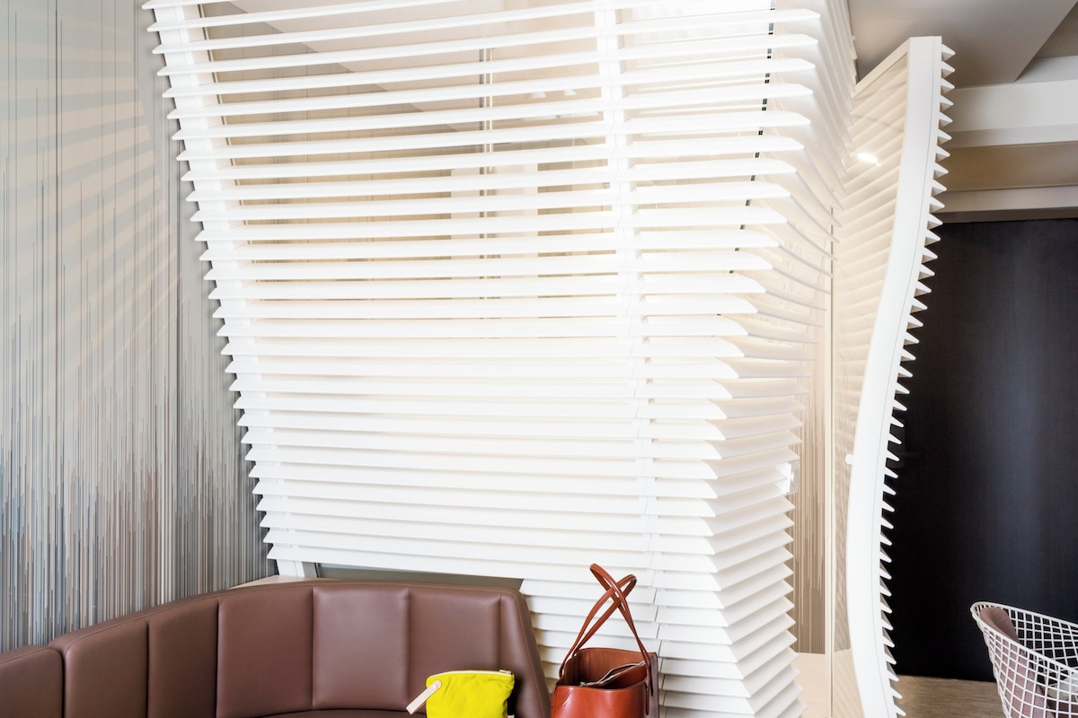 Une chambre d’hôtel moderne avec un canapé marron aux lignes courbes, un petit sac jaune, un sac en cuir marron et une chaise blanche.