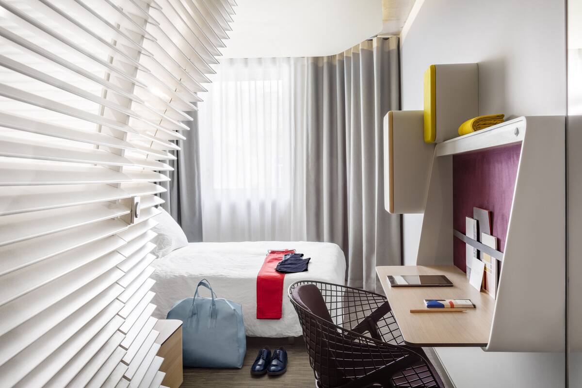 Une chambre d’hôtel moderne équipée d’un lit, d’un bureau, d’une chaise et d’une grande fenêtre avec des stores.