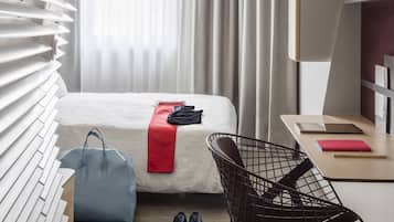 A modern hotel room with a bed, a desk, a chair, and a window with blinds.