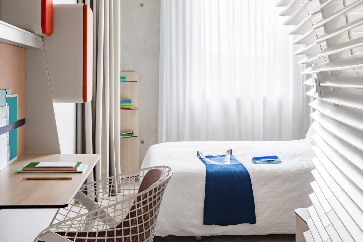 Une chambre d’hôtel moderne équipée d’un lit, d’un bureau, d’une chaise et d’une fenêtre avec des stores.