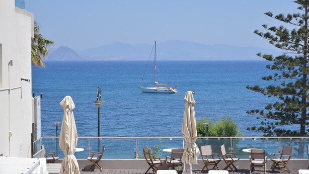 Eine Terrasse mit Außenplätzen mit Blick auf das Meer, ein Segelboot in der Ferne und Berge im Hintergrund.