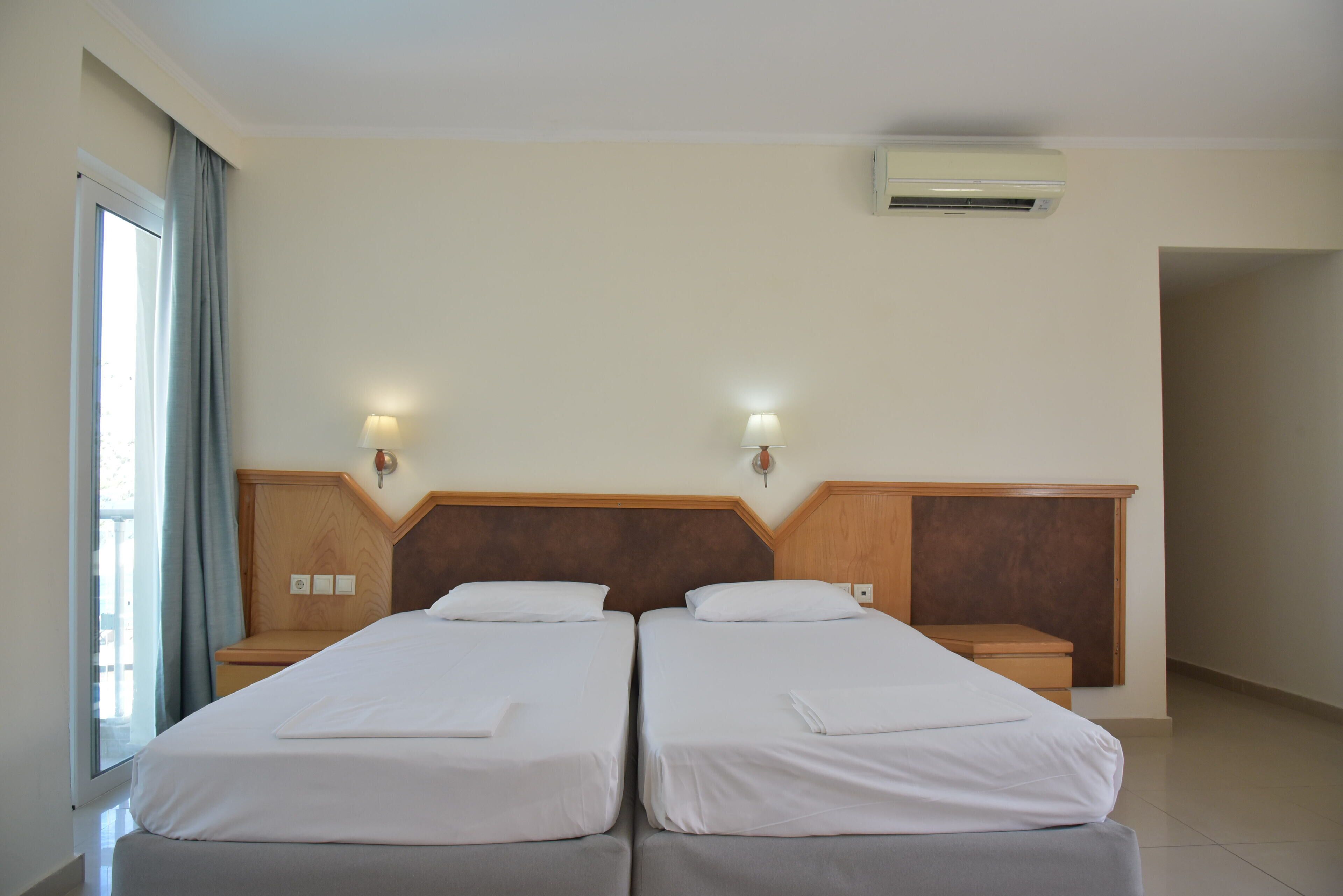 Une chambre d’hôtel avec deux lits simples, une tête de lit en bois, des lampes de chevet, un appareil de climatisation et une fenêtre avec des rideaux.