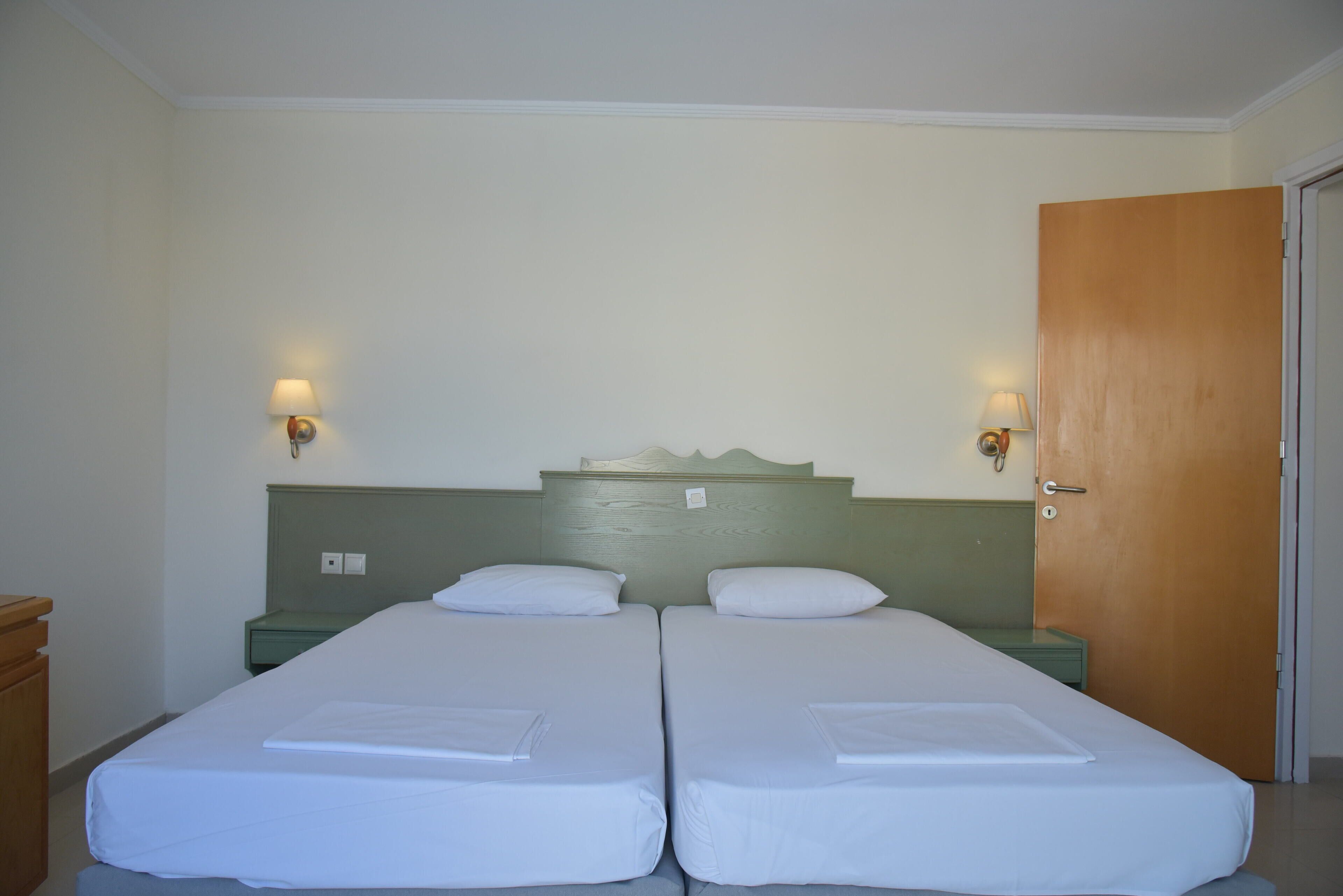 A room with two single beds, each with a white mattress and pillow, a wooden headboard, and a green nightstand with a lamp.
