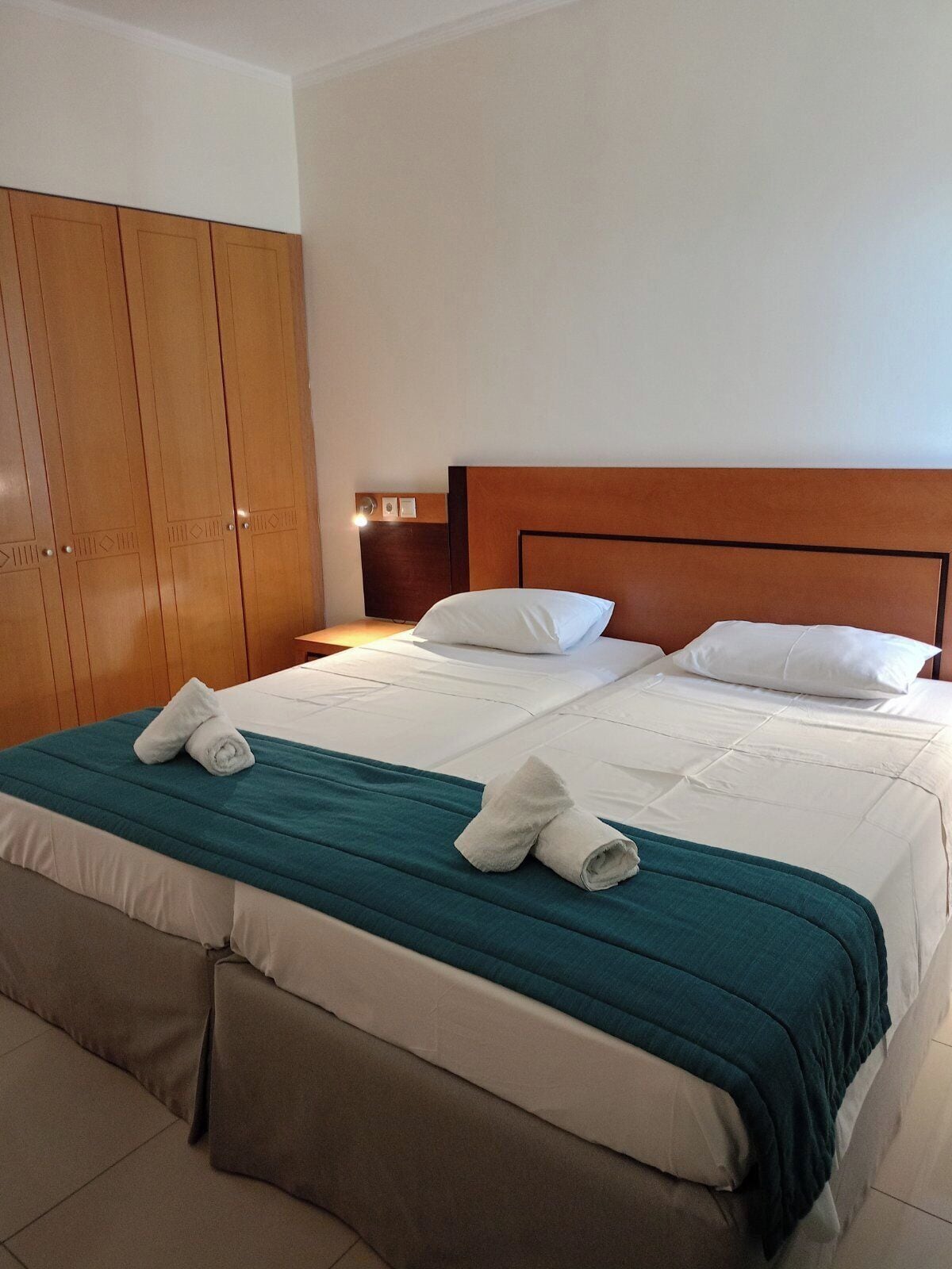 Habitación de hotel con una cama grande, dos almohadas, un armario de madera y una mesita de noche con lámpara.