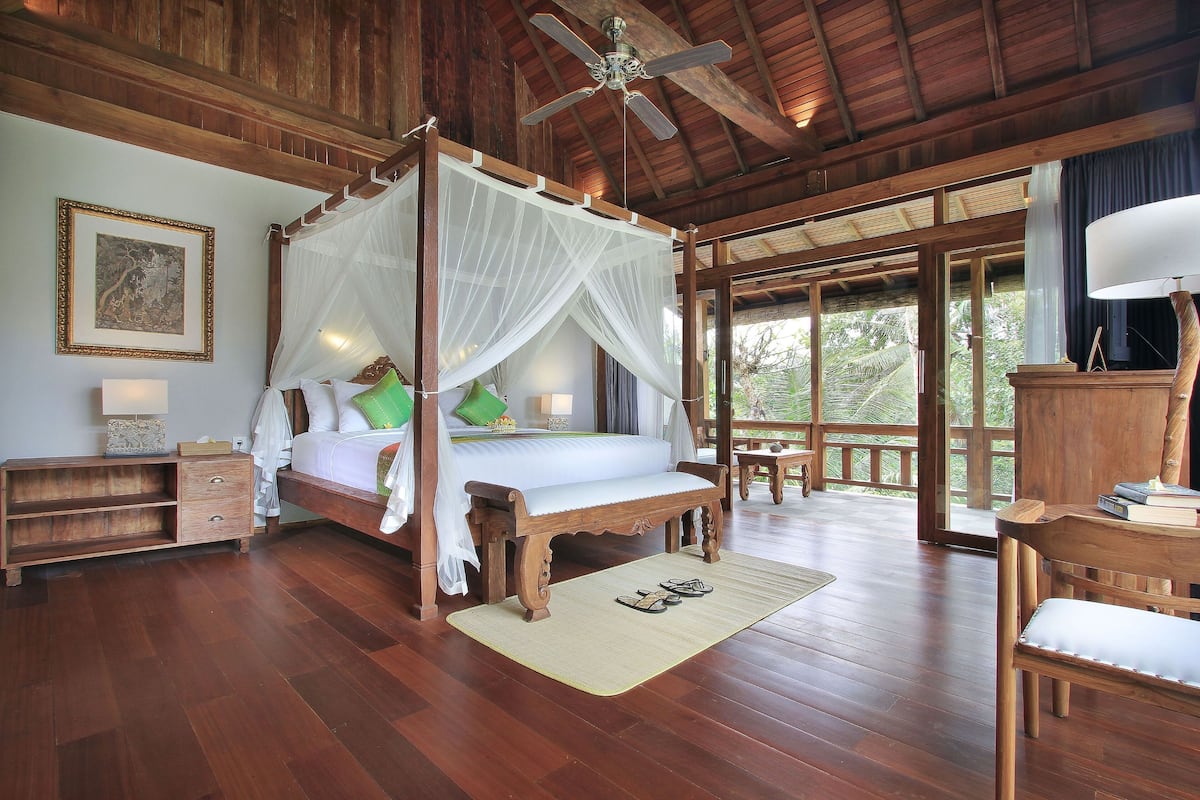 A wooden four-poster bed with a canopy, a wooden nightstand, a bedside table with a lamp, and a view of the outdoors through large windows.