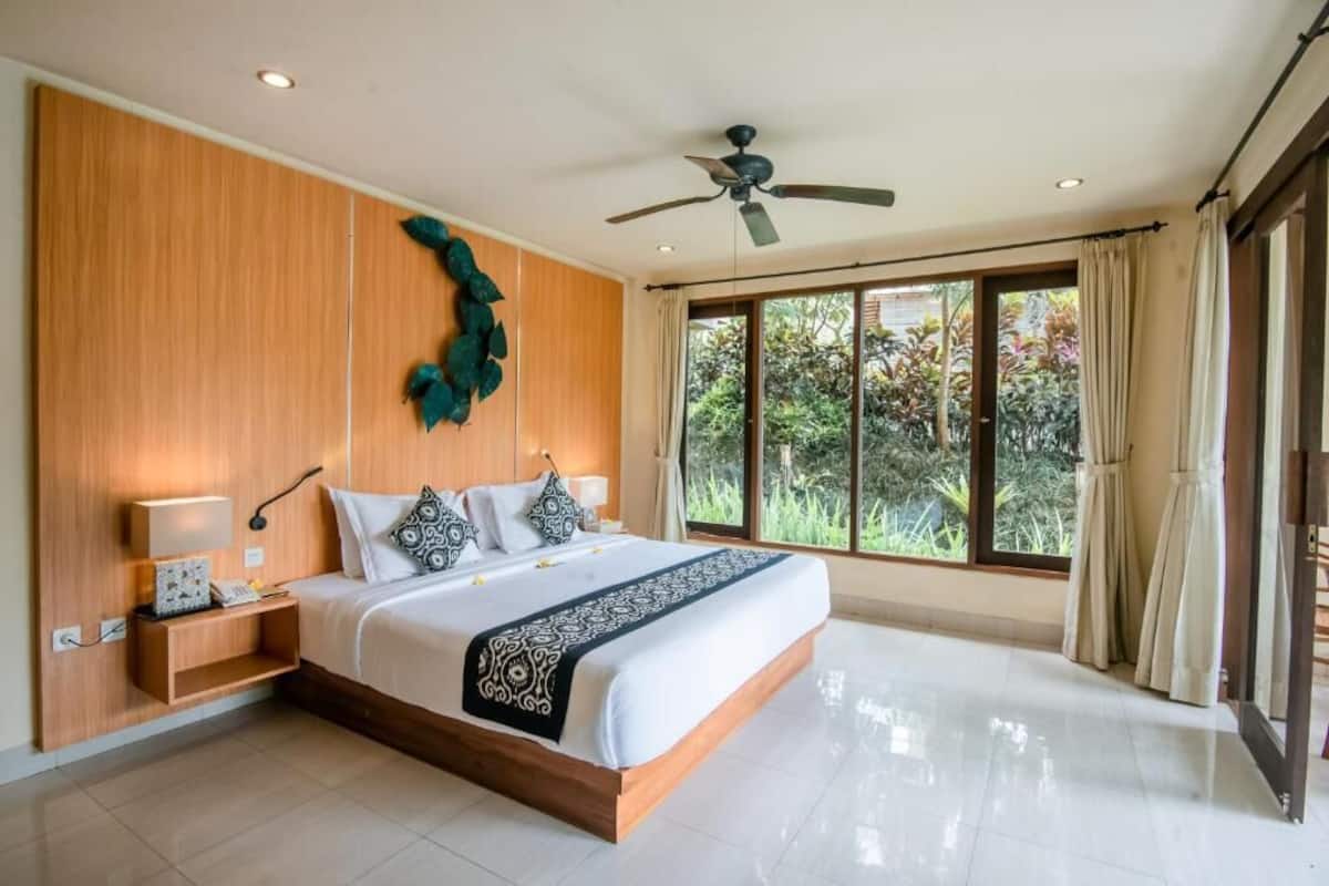 A large bed with a wooden headboard, a ceiling fan, and a view of greenery through large windows.