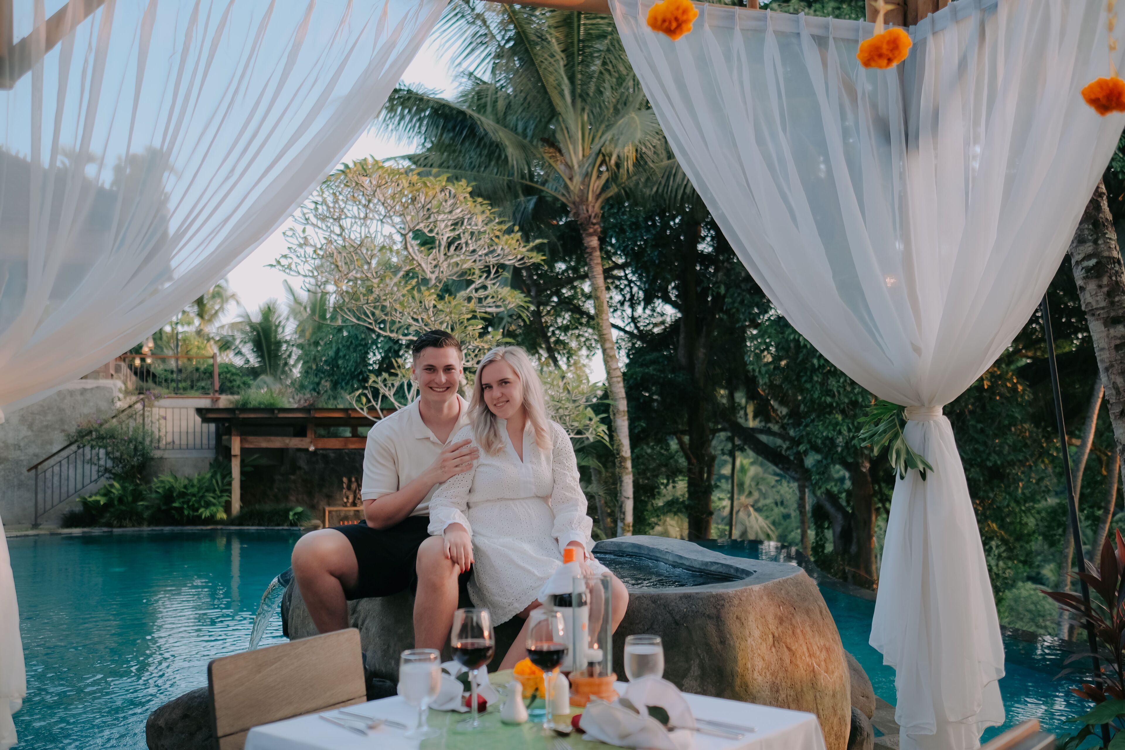 Una pareja sentada en una mesa junto a la piscina, con bebidas y una botella, rodeada de cortinas blancas y palmeras.