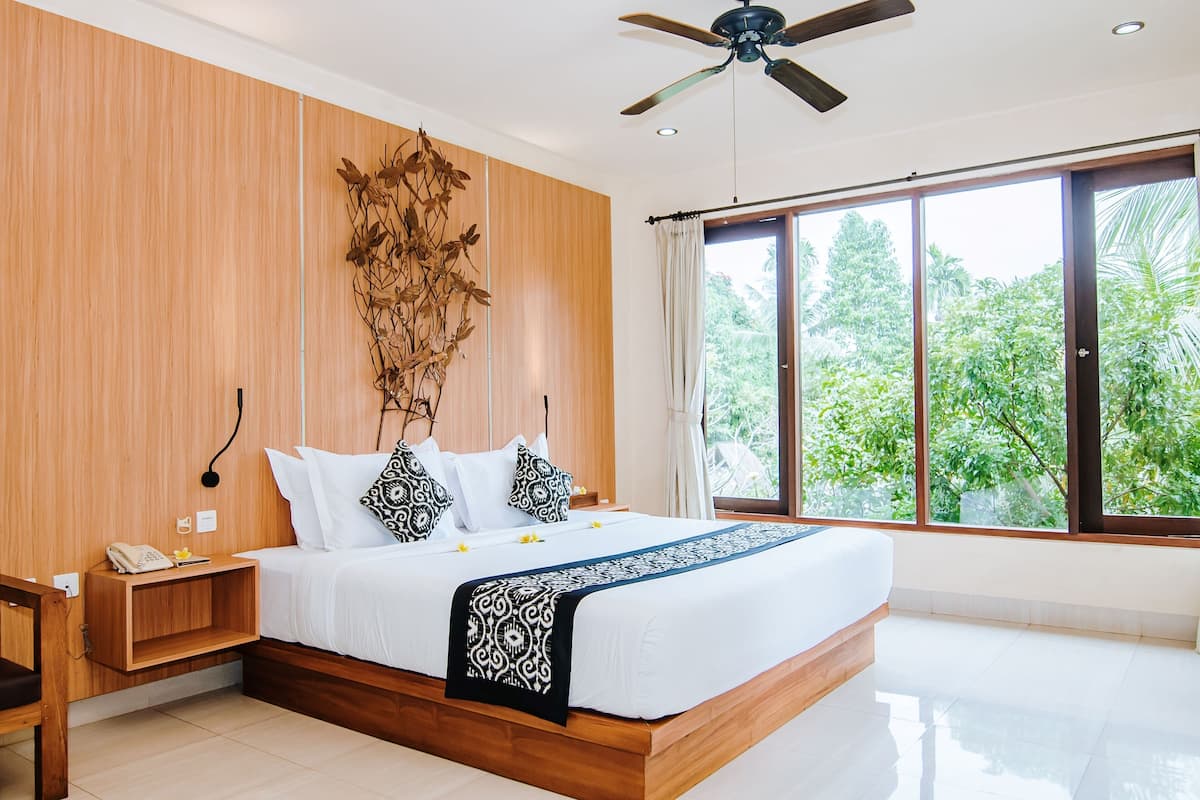 A modern bedroom with a wooden headboard, a large bed with white bedding, a bedside table with a phone, and a view of greenery through large windows.