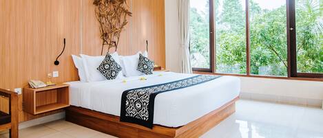 A modern bedroom with a wooden headboard, a large bed with white bedding, a bedside table with a phone, and a view of greenery through large windows.