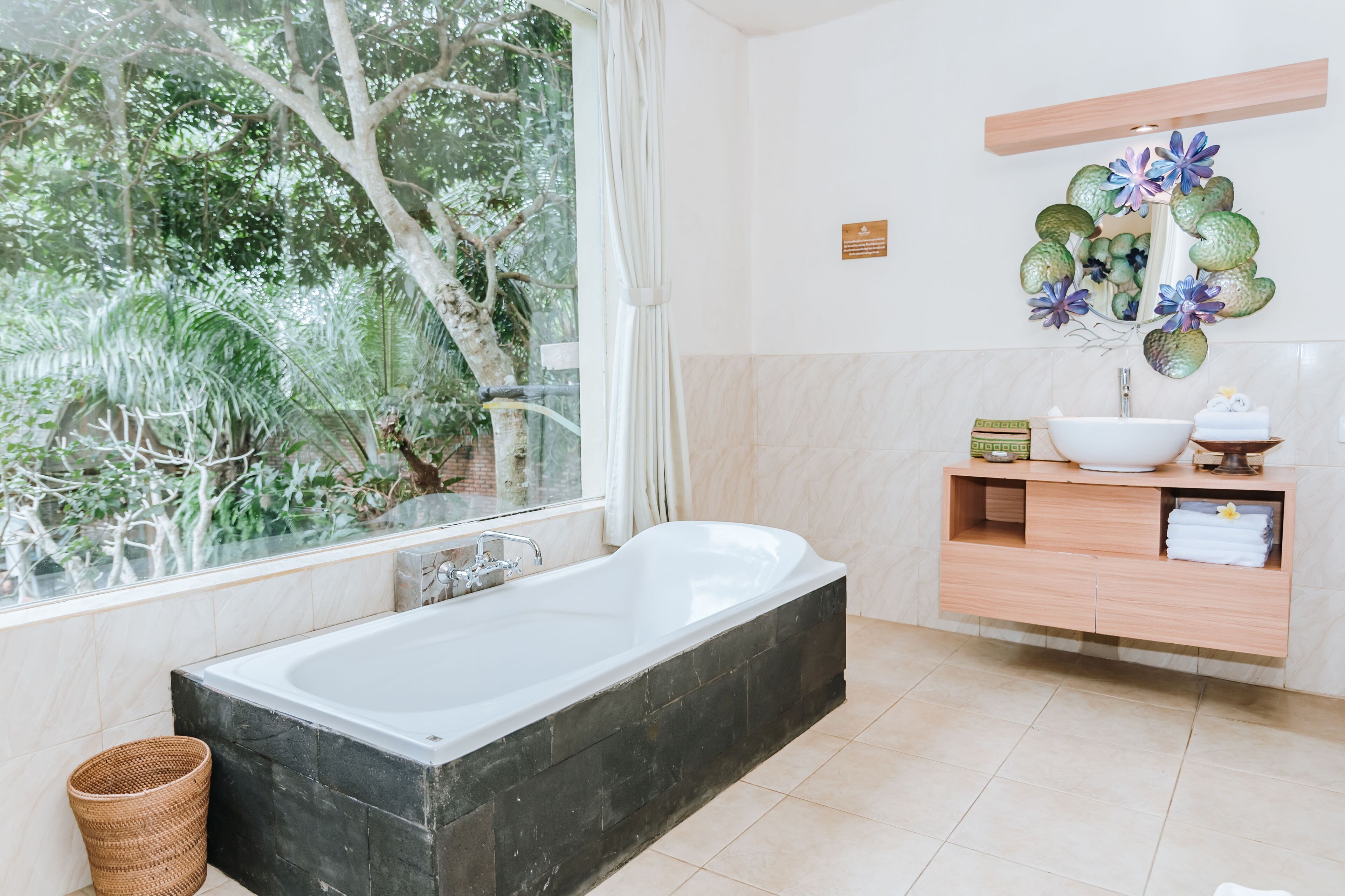 A modern bathroom with a bathtub, a large window with a view of greenery, a wooden vanity with a bowl sink, and a mirror with decorative flowers.