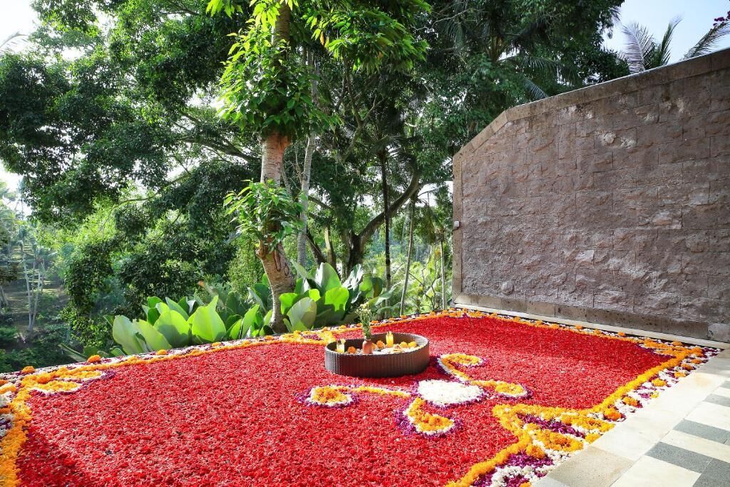 A garden area with a red flower carpet, a central decorative fire pit, and lush greenery in the background.