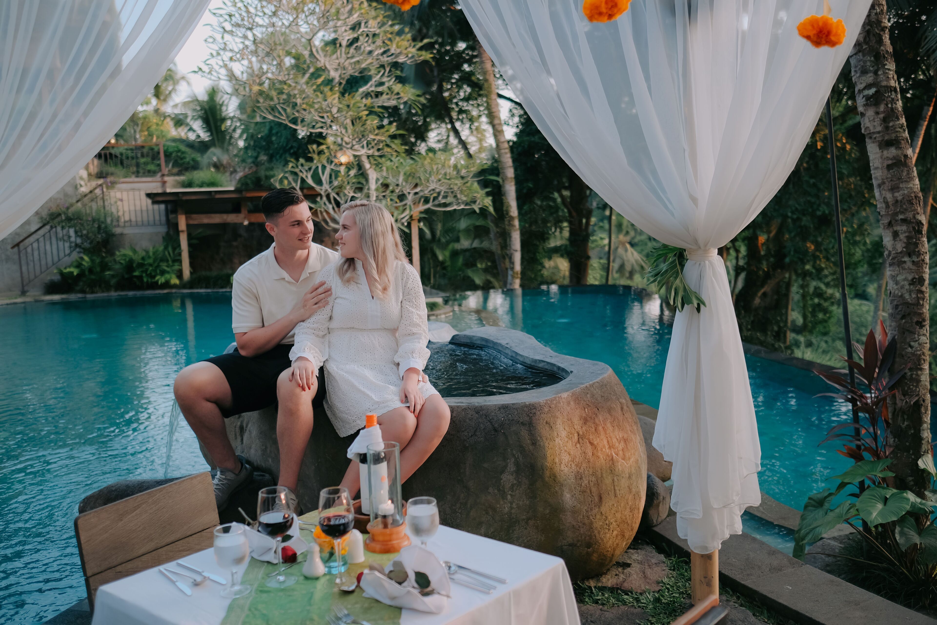 A couple sitting by a poolside table with drinks and a candle, surrounded by greenery and a clear pool.