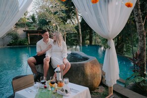 A couple sitting by a poolside table with drinks and a candle, surrounded by greenery and a clear pool. - Bucu View Ubud Resort (Ubud)