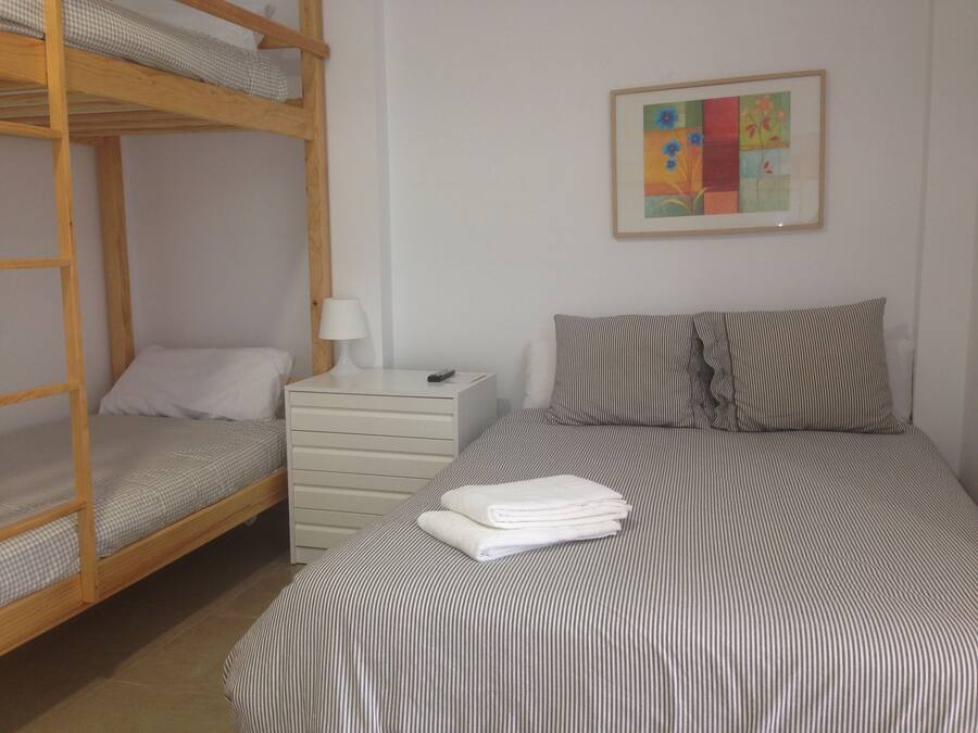 Een slaapkamer met een stapelbed, een eenpersoonsbed, een nachtkastje, een lamp en een lijst aan de muur.