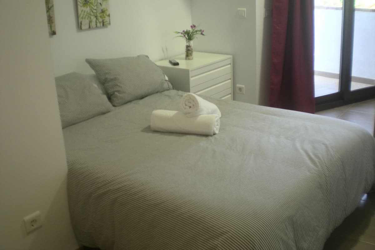 A neatly made bed with a rolled towel, a bedside table with a vase of flowers, and a window with curtains.