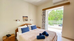 A bedroom with a bed, a nightstand, a window with curtains, and a view of greenery outside. - Angela Hotel and Apartments (Corfu)
