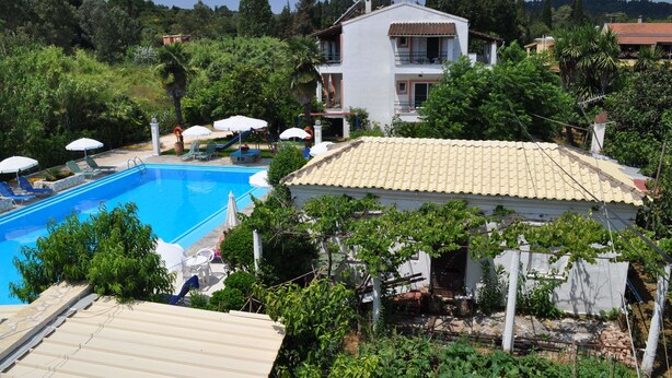 A pool area with lounge chairs and umbrellas, surrounded by greenery and a building with a tiled roof. - Angela Hotel and Apartments (Corfu)