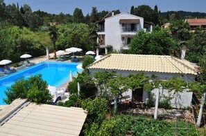 A pool area with lounge chairs and umbrellas, surrounded by greenery and a building with a tiled roof. - Angela Hotel and Apartments (Corfu)