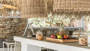 A rustic outdoor dining area with a stone wall backdrop, a wooden table with fruits and cookies, and wicker hanging lights. - Verina Terra (Sifnos)