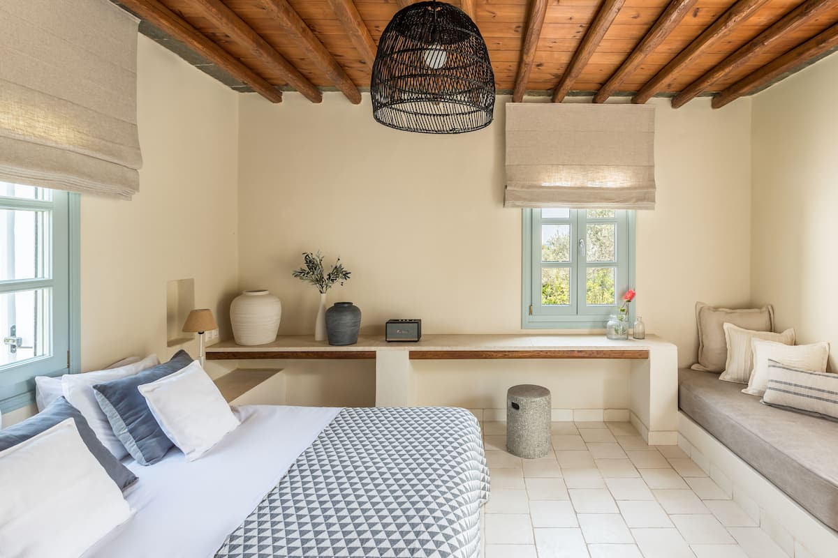 Un dormitorio con cama, mesita de noche, lámpara, jarrón y ventana con vistas.