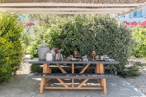 An outdoor dining area with a wooden table and benches, surrounded by lush greenery and a stone pathway. - Verina Terra (Sifnos)