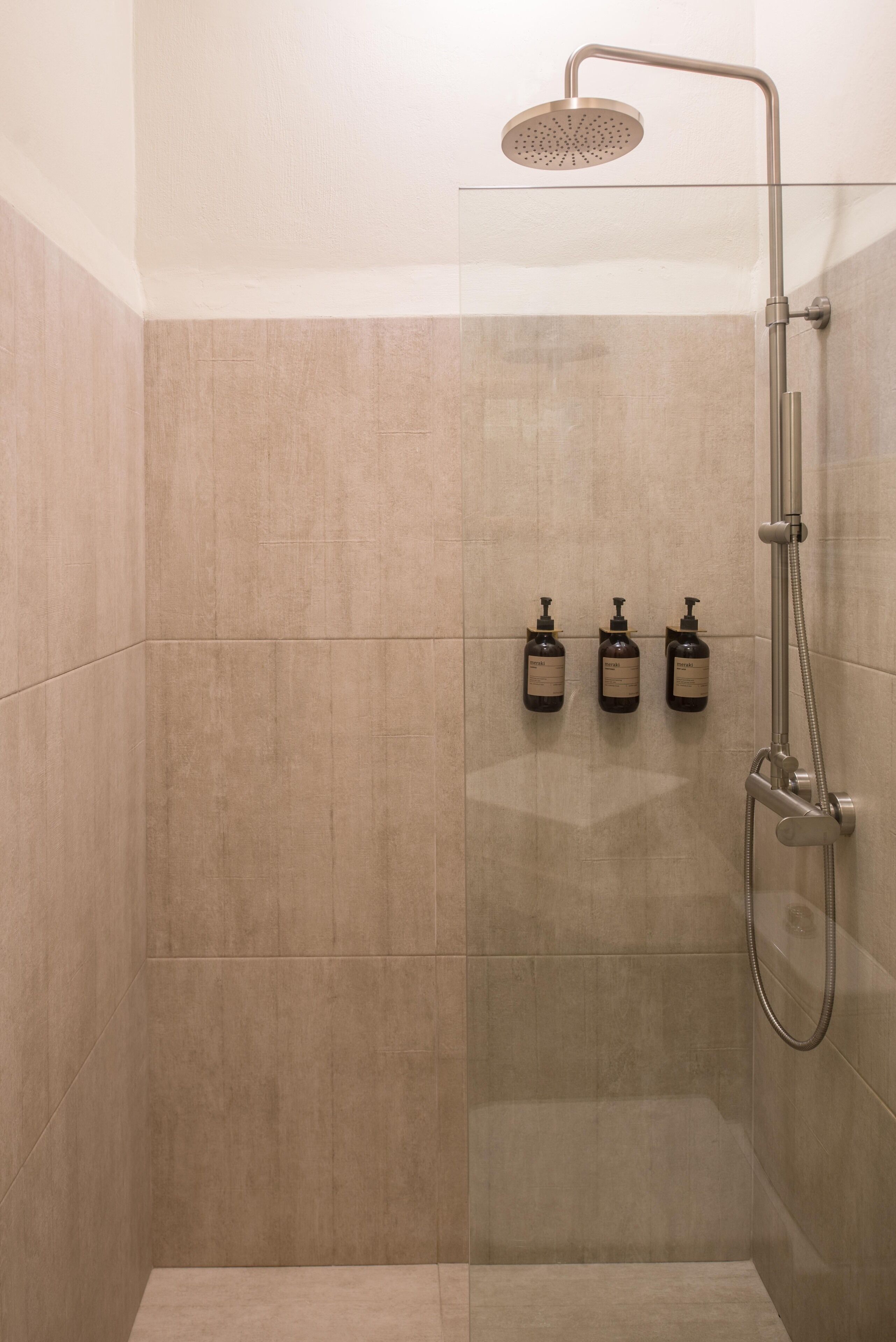 A modern shower with a glass enclosure, a handheld showerhead, and three bottles of shampoo on the wall.