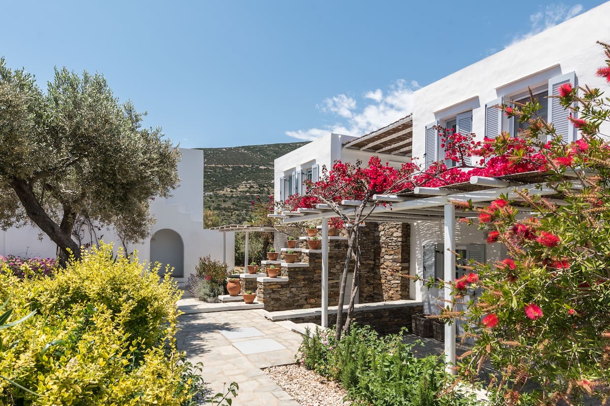 Un edificio blanco de fachada de piedra, una pérgola con flores rojas y un camino que lleva a una entrada en arco.