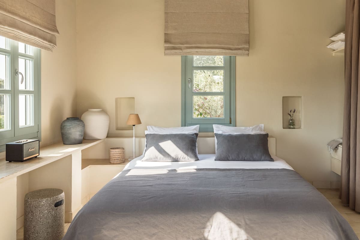 Un dormitorio con una cama, una ventana, una lámpara y una jarra en la mesita de noche.