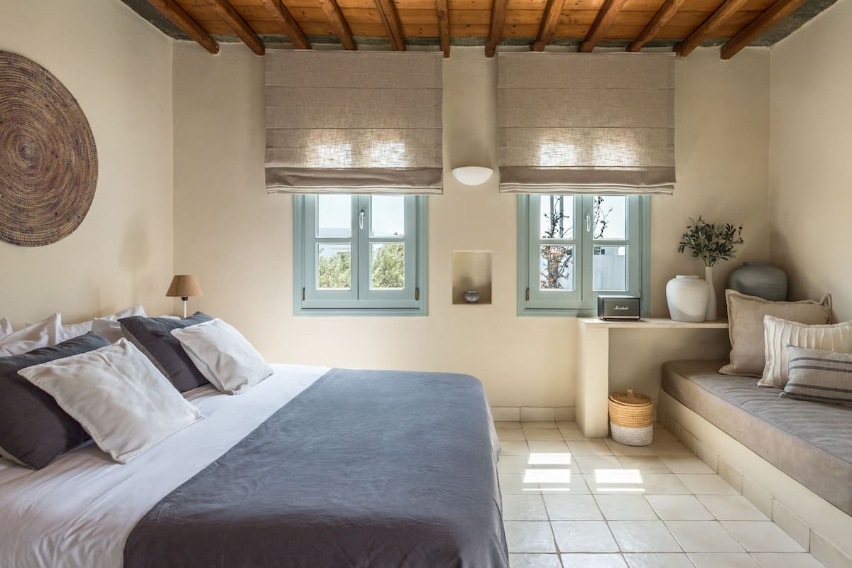 Un dormitorio con una cama grande, dos ventanas con marcos azules, una mesita de noche con lámpara y una pequeña zona de estar con cojines.