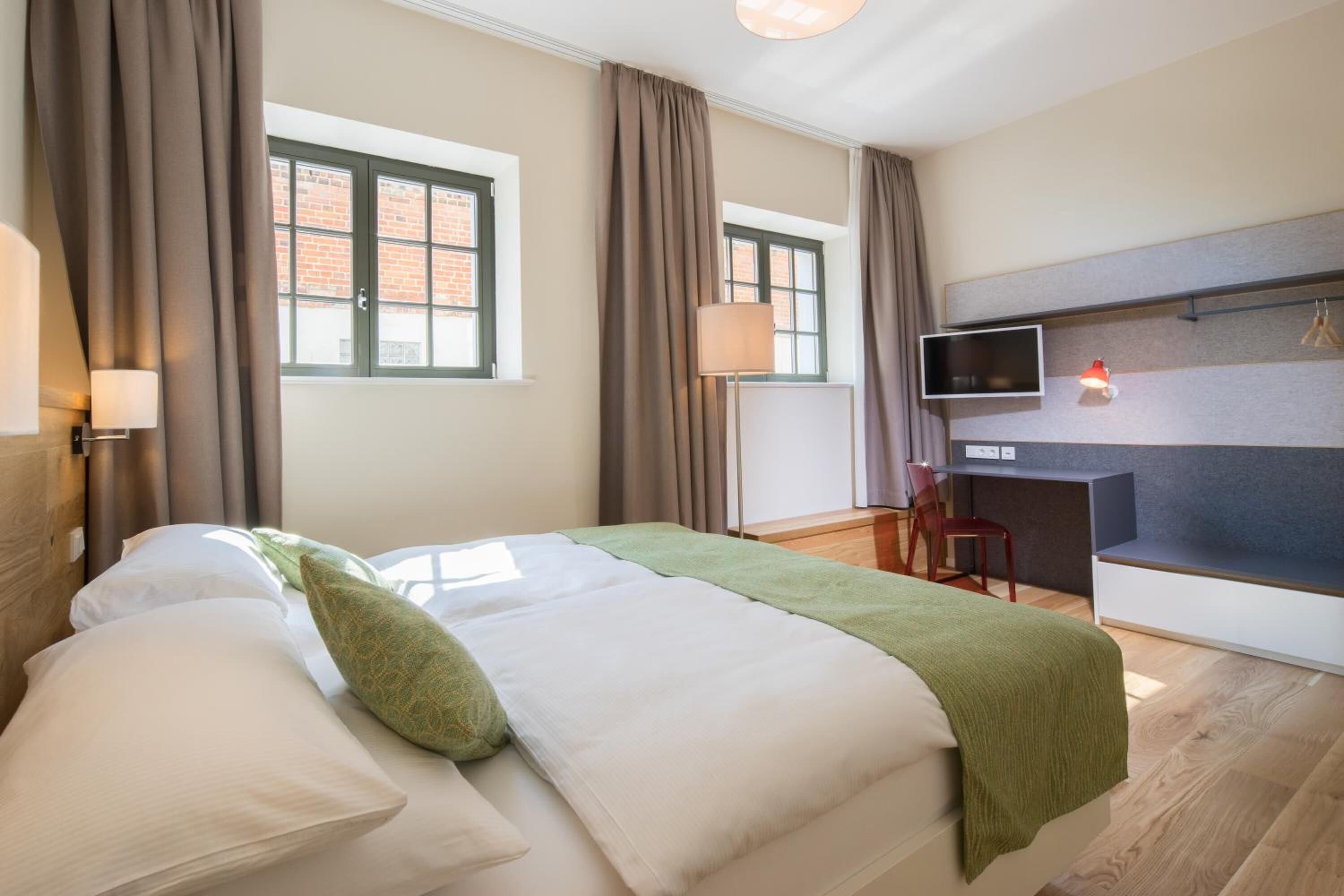 Una habitación de hotel con cama, escritorio, silla, televisor y ventana con cortinas.