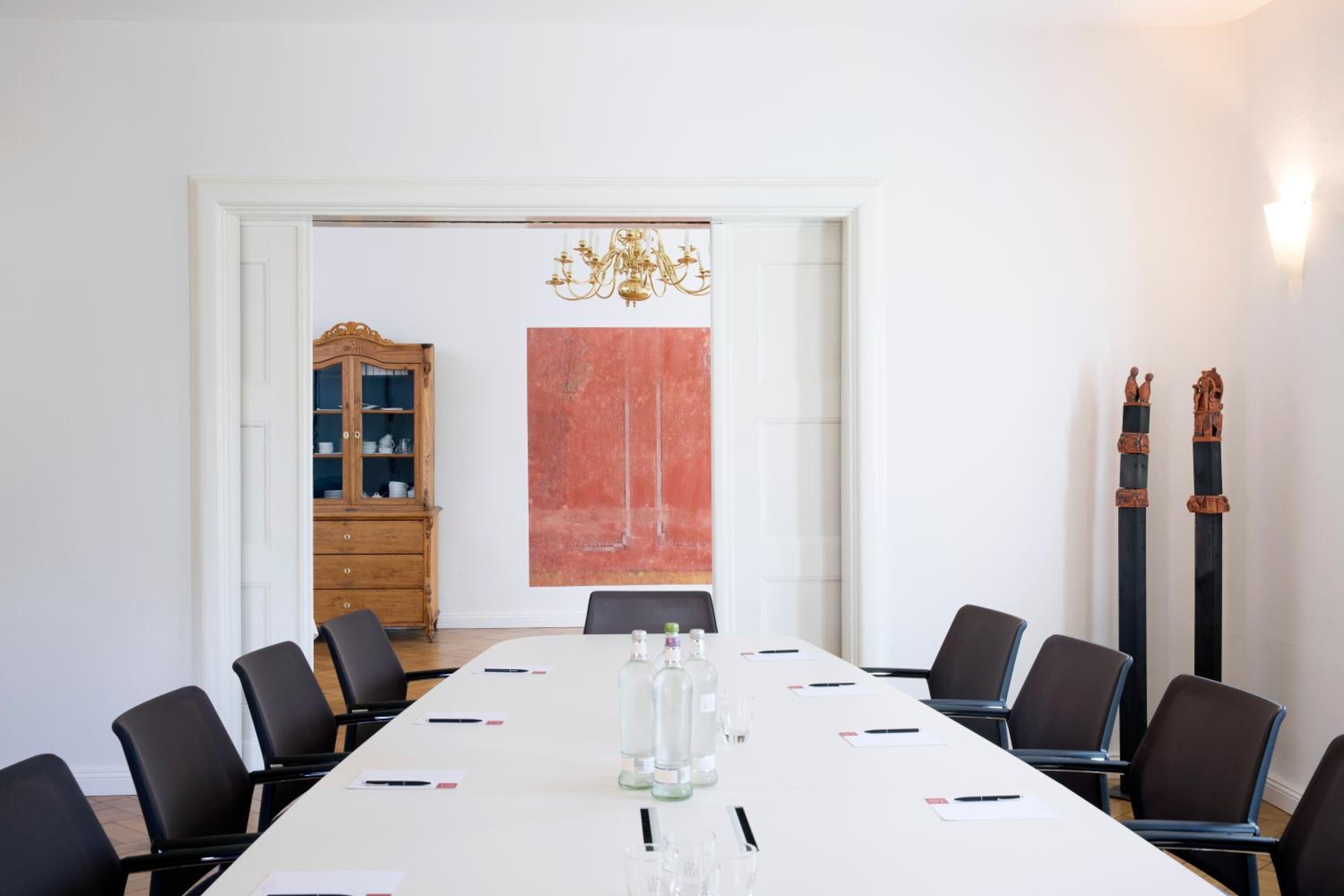Un salón de conferencias con una mesa larga, sillas negras, un mueble de madera y un cuadro abstracto rojo.