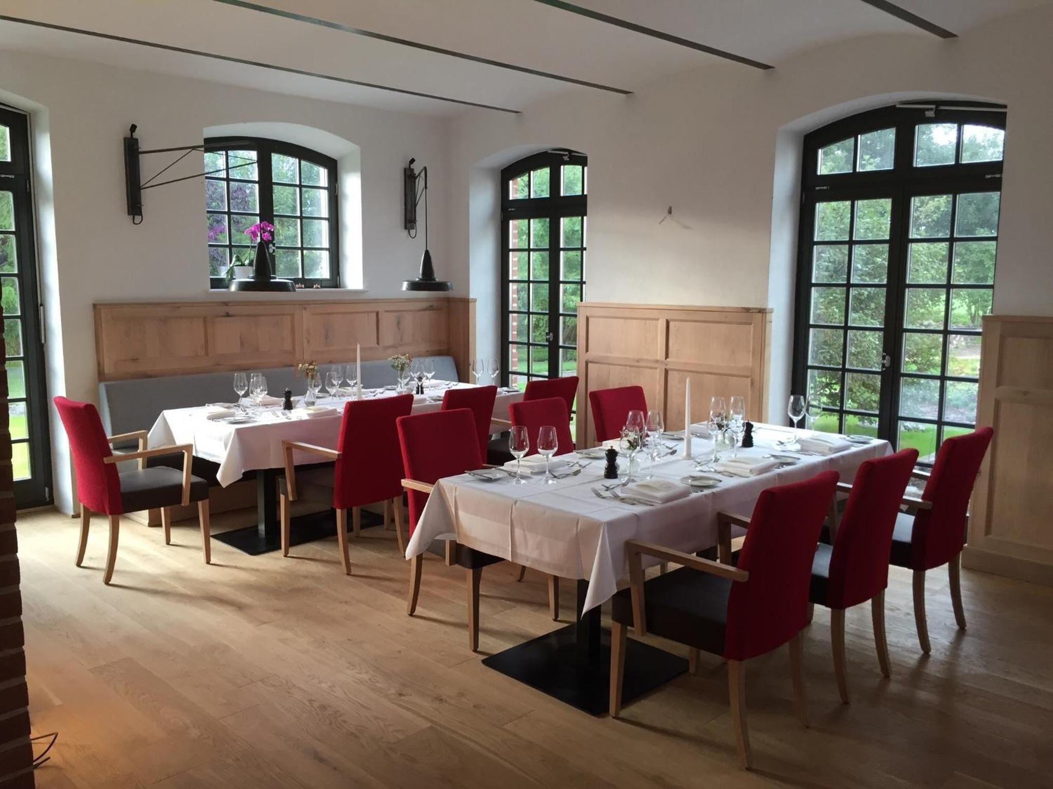 Comedor con varias mesas preparadas para una comida, suelo de madera y ventanas amplias con vistas a zonas verdes.
