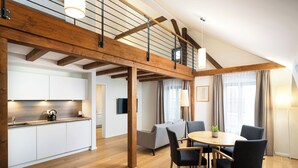Ein geräumiger Wohnbereich mit hoher Decke, Holzbalken, einem Esstisch mit Stühlen, einer Couch und einer Küchenzeile mit weißen Schränken und einer Holz Arbeitsplatte.