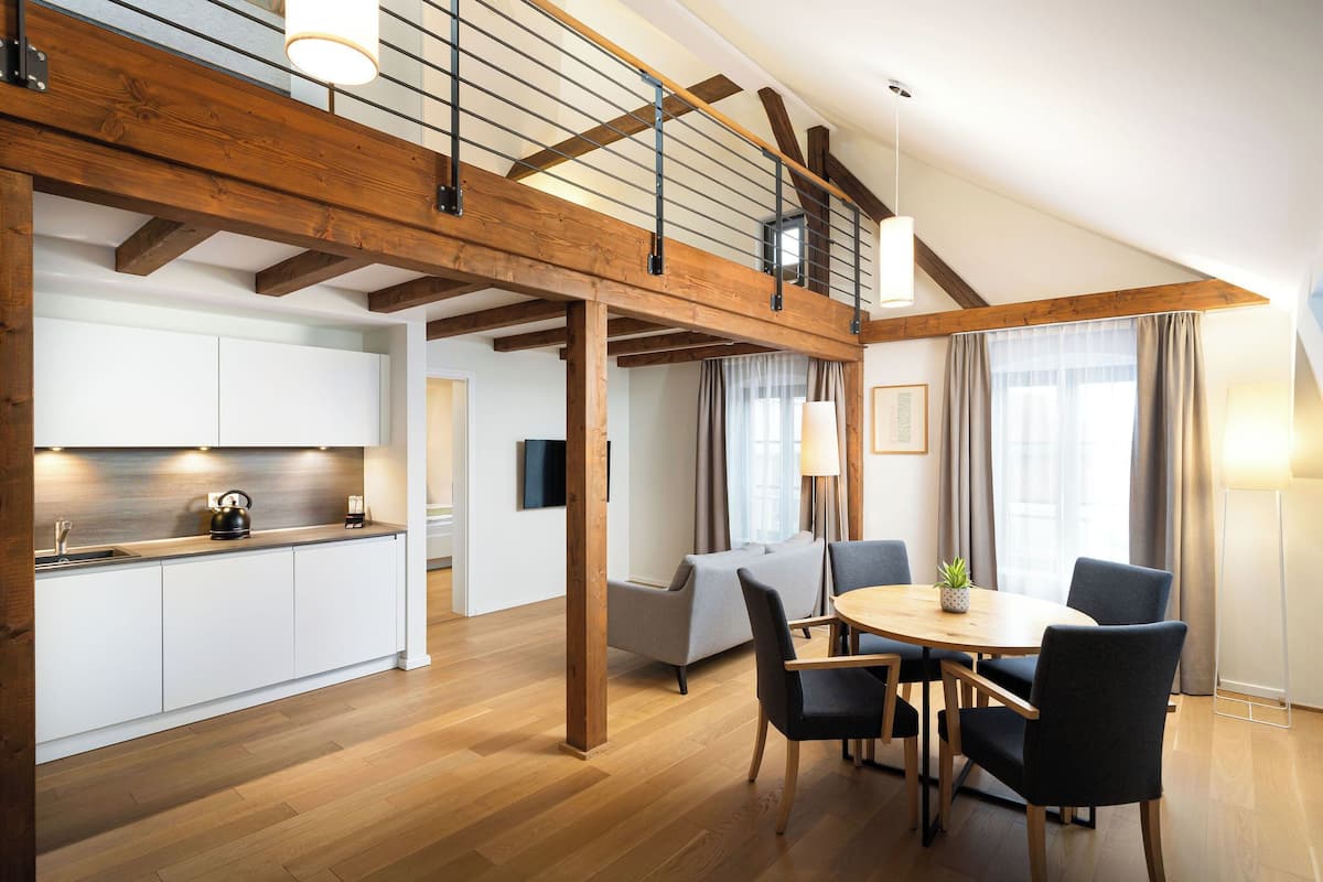 Un salón espacioso con techo alto, vigas de madera, una mesa de comedor con sillas, un sofá y una zona de cocina con armarios blancos y encimera de madera.