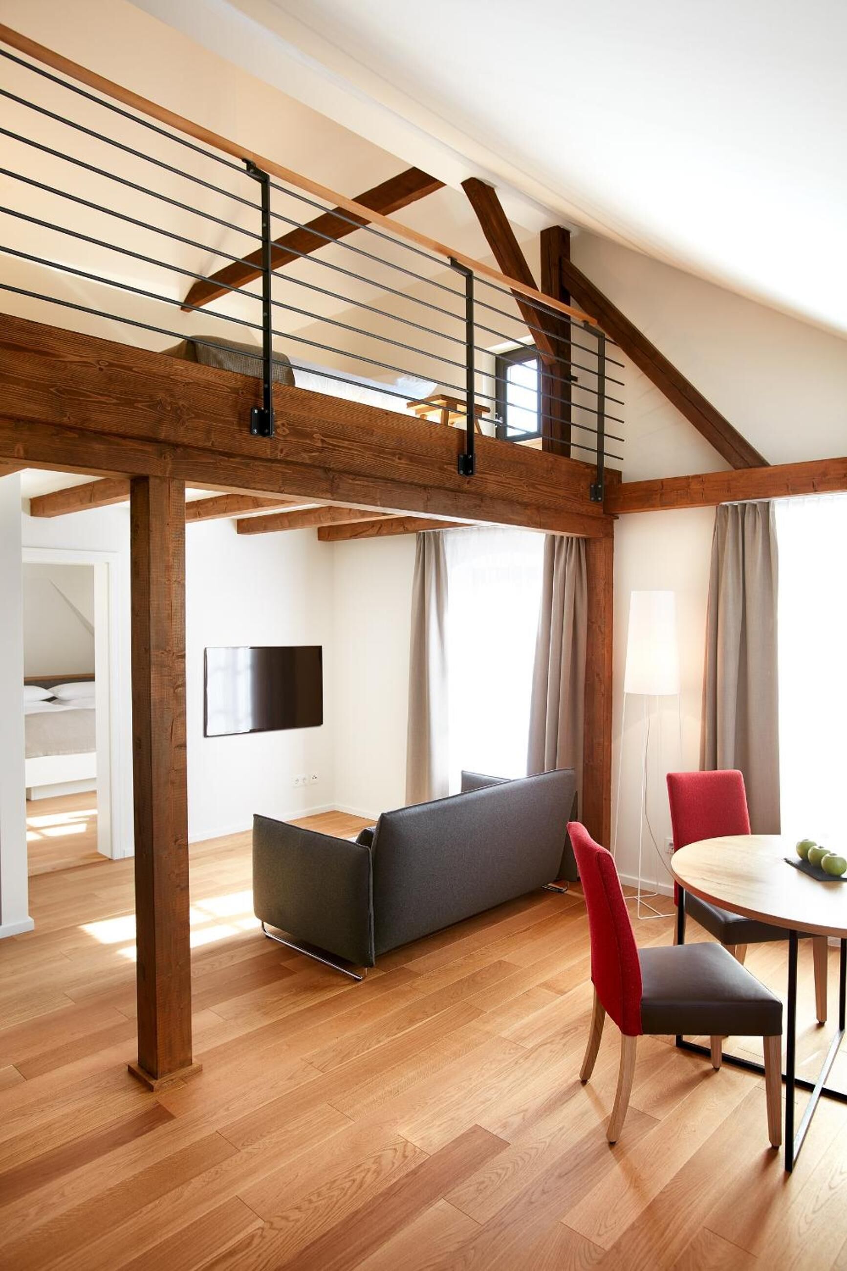 Una habitación moderna estilo loft con vigas de madera expuestas, una zona de estar en altura, una mesa de comedor con sillas y un televisor de pantalla plana montado en la pared.