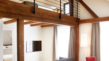 Una habitación moderna estilo loft con vigas de madera expuestas, una zona de estar en altura, una mesa de comedor con sillas y un televisor de pantalla plana montado en la pared.