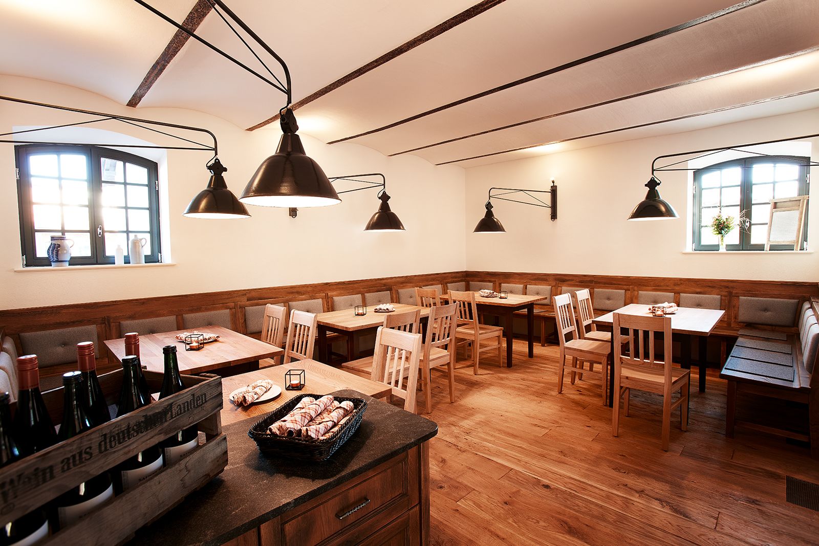 A restaurant with wooden tables and benches, pendant lighting, and a wooden bar area with bottles.
