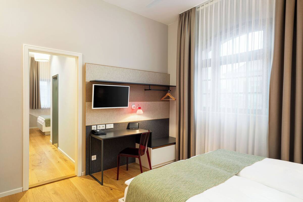 Habitación de hotel con cama, escritorio y silla, televisor y ventana con cortinas.