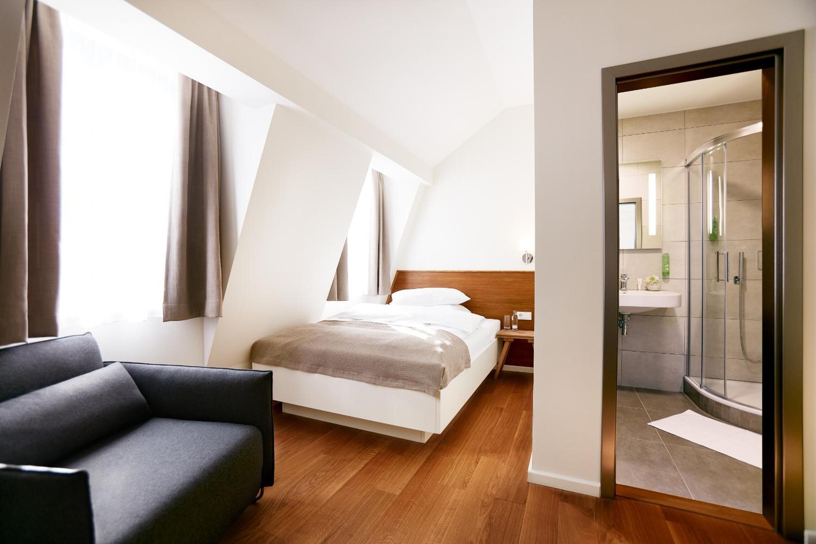 A modern hotel room with a bed, a nightstand, a sofa, and a shower area.